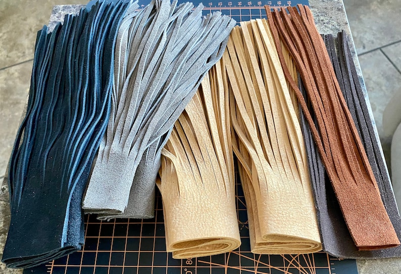 May include: Five strips of leather fringe in various colors, including black, gray, beige, brown, and dark brown. The fringe is cut into long, thin strips and is arranged in a row on a black and orange cutting mat.