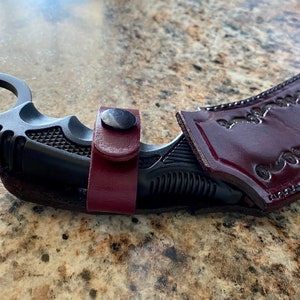 Puede incluir: Un cuchillo karambit negro con un anillo para el dedo y un mango texturizado, asegurado en una funda de cuero burdeos. La funda presenta detalles decorativos en relieve y un cierre a presi&oacute;n. El cuchillo est&aacute; dise&ntilde;ado para la autodefensa.
