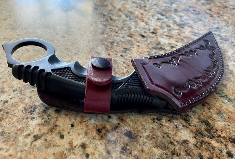 Puede incluir: Un cuchillo karambit negro con un anillo para el dedo y un mango texturizado, asegurado en una funda de cuero burdeos. La funda presenta detalles decorativos en relieve y un cierre a presi&oacute;n. El cuchillo est&aacute; dise&ntilde;ado para la autodefensa.