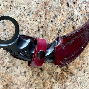 Puede incluir: Un cuchillo karambit negro con un anillo para el dedo y una funda de cuero burdeos. El cuchillo tiene una hoja curva y un mango texturizado. La funda est&aacute; decorada con un dise&ntilde;o floral en relieve y un cierre a presi&oacute;n.