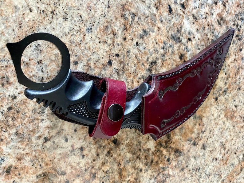 Puede incluir: Un cuchillo karambit negro con un anillo para el dedo y una funda de cuero burdeos. El cuchillo tiene una hoja curva y un mango texturizado. La funda est&aacute; decorada con un dise&ntilde;o floral en relieve y un cierre a presi&oacute;n.