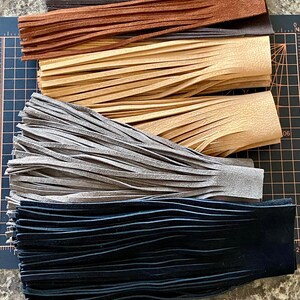 May include: Four strips of leather fringe in different colors: black, gray, beige, and brown. The fringe is cut into thin strips and is arranged in a stack.
