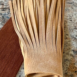 May include: A close-up of a light brown leather fringe. The fringe is folded in half and the ends are visible. The fringe is made of many thin strips of leather.