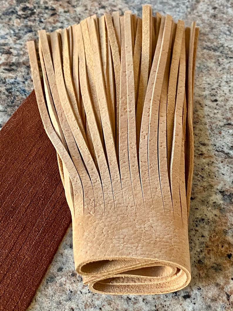 May include: A close-up of a light brown leather fringe. The fringe is folded in half and the ends are visible. The fringe is made of many thin strips of leather.