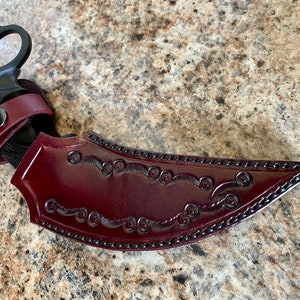 Puede incluir: Un cuchillo karambit negro con una hoja curva y un anillo para el dedo. El cuchillo se combina con una funda de cuero burdeos con adornos decorativos y una correa de cuero a juego.