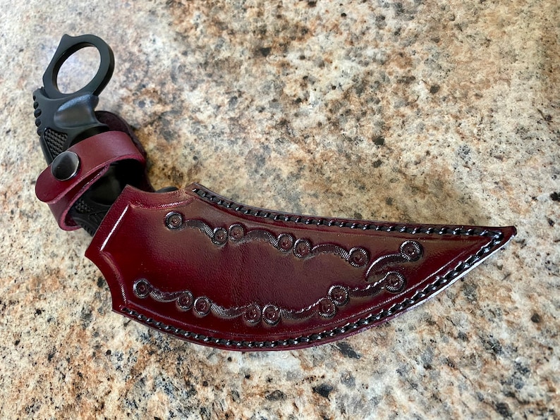 Puede incluir: Un cuchillo karambit negro con una hoja curva y un anillo para el dedo. El cuchillo se combina con una funda de cuero burdeos con adornos decorativos y una correa de cuero a juego.