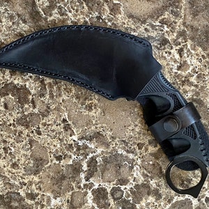 May include: Black leather sheath with a black karambit knife. The knife has a finger ring and a textured handle.