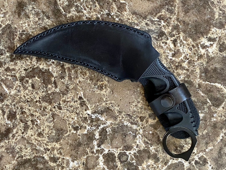 May include: Black leather sheath with a black karambit knife. The knife has a finger ring and a textured handle.