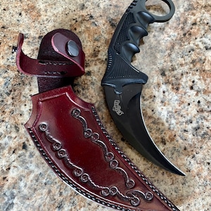Puede incluir: Un cuchillo karambit negro con una hoja curva y un anillo para el dedo, junto a una funda de cuero burdeos. La funda tiene adornos decorativos y un cierre a presi&oacute;n. El mango del cuchillo tiene un agarre texturizado.
