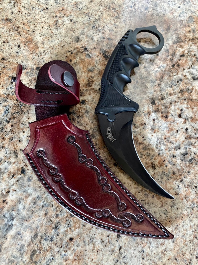 Puede incluir: Un cuchillo karambit negro con una hoja curva y un anillo para el dedo, junto a una funda de cuero burdeos. La funda tiene adornos decorativos y un cierre a presi&oacute;n. El mango del cuchillo tiene un agarre texturizado.