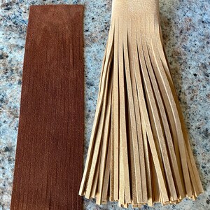 May include: Two strips of leather fringe, one brown and one tan. The brown fringe is flat and the tan fringe is gathered into a bunch.