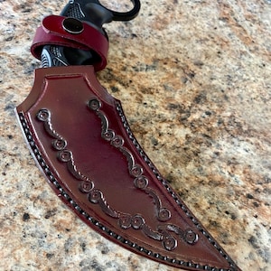 Puede incluir: Un cuchillo karambit negro con una funda y correa de cuero burdeos. La funda presenta adornos decorativos y un borde cosido. El mango tiene un anillo para un agarre seguro.