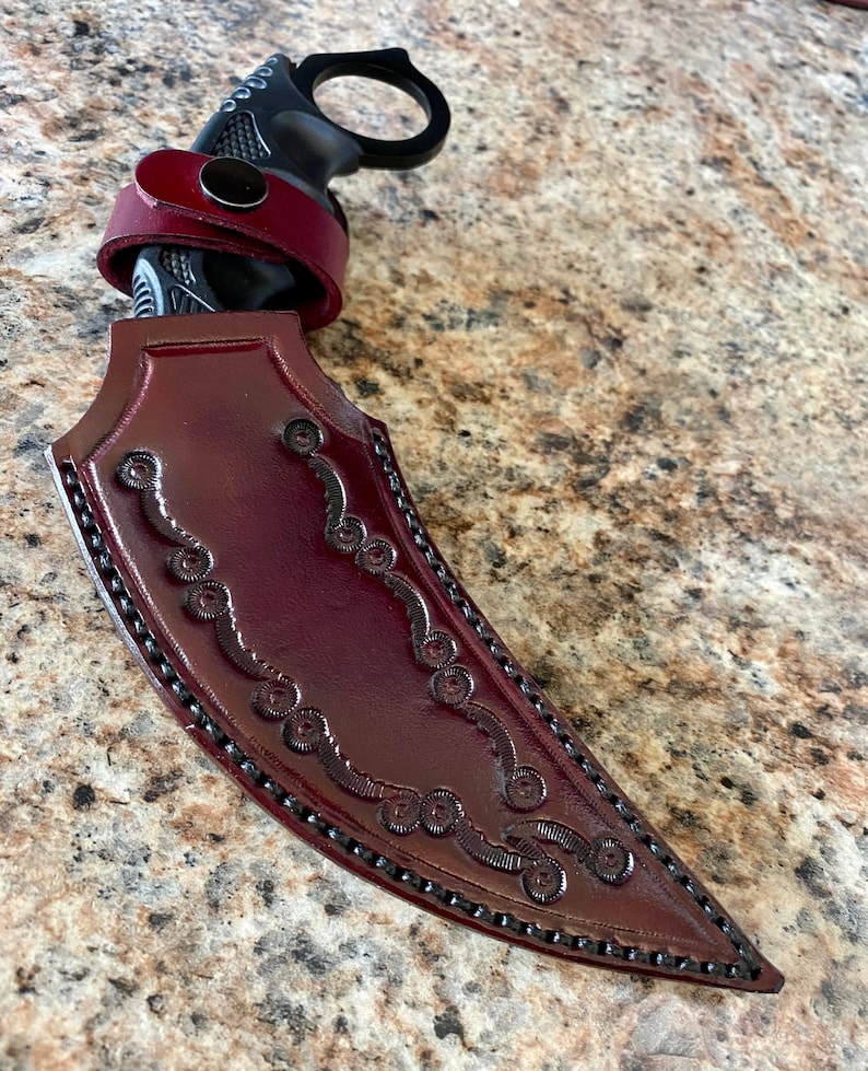 Puede incluir: Un cuchillo karambit negro con una funda y correa de cuero burdeos. La funda presenta adornos decorativos y un borde cosido. El mango tiene un anillo para un agarre seguro.