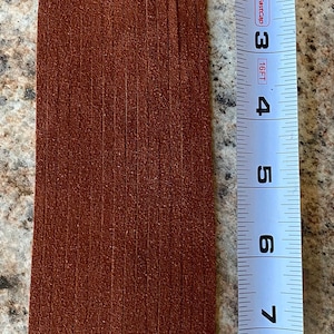 May include: A brown leather fringe strip measuring 9 inches long. The fringe is made of thin strips of leather that are evenly spaced and hang down from a rectangular piece of leather.