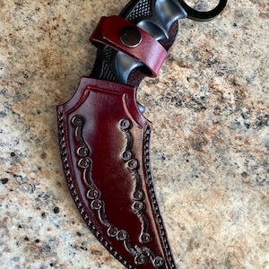 May include: A black karambit knife with a finger ring, set in a burgundy leather sheath. The sheath has decorative stitching and a leather strap with a snap closure. The knife's handle is black and textured.