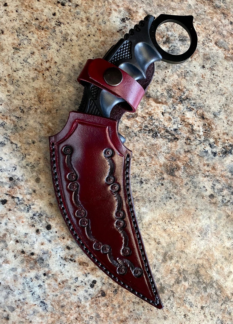 Puede incluir: Un cuchillo karambit negro con un anillo para el dedo, dentro de una funda de cuero burdeos. La funda tiene costuras decorativas y una correa de cuero con cierre de bot&oacute;n a presi&oacute;n. El mango del cuchillo es negro y texturizado.