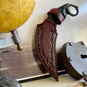 Puede incluir: Un cuchillo karambit con una funda de cuero burdeos. El cuchillo tiene un mango negro y un anillo para el dedo. La funda est&aacute; decorada con un dise&ntilde;o en relieve. Un globo terr&aacute;queo antiguo y un candado oxidado est&aacute;n en el fondo.