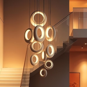 May include: A contemporary pendant light fixture featuring multiple white, ring-shaped lights suspended from the ceiling. The illuminated lights create a warm ambiance in a modern interior, with a staircase and a neutral colour scheme.