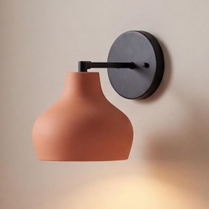 May include: A wall-mounted sconce featuring a matte black circular base and a horizontal arm supporting a bell-shaped terracotta-coloured lampshade. The fixture emits a warm downward light, illuminating a small ceramic bowl resting on stacked books.