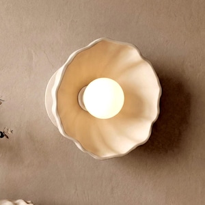Può includere: Applique da parete bianco e beige con un design unico a forma di fiore. L'apparecchio presenta una lampadina centrale rotonda e luminosa, circondata da un paralume a forma di petalo smerlato. Montato su una parete di colore neutro.