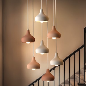 May include: A collection of pendant lights in white and terracotta colours hang from the ceiling. The lights are suspended at different heights, illuminating a staircase. The design is modern, with a minimalist aesthetic.