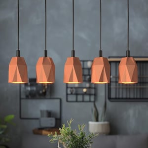 May include: A set of five hanging pendant lights featuring geometric, faceted terracotta-coloured shades suspended by thin black cords. Each light fixture emits a warm glow from the bottom opening, showcasing a structured, angular design.