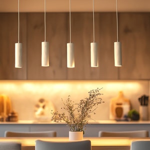 May include: Five white cylindrical pendant lights suspended above a wooden table. The lights are evenly spaced, illuminating the dining area. A vase of dried flowers sits on the table. The background features wooden cabinets and a kitchen setup.