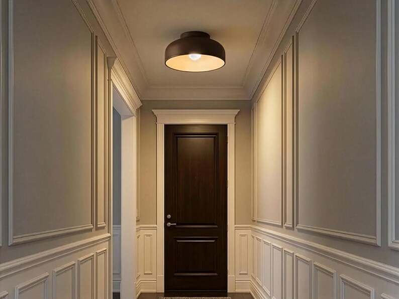 A hallway with light gray walls and white trim. A dark brown door is at the end of the hallway. A brown, circular ceiling light with a yellow glow hangs from the ceiling.