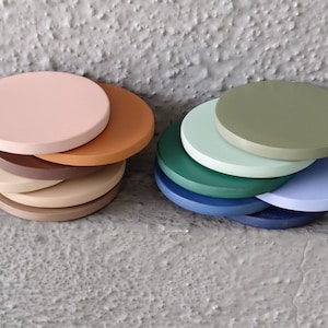 Könnte beinhalten: Zwei Stapel runde, bemalte Untersetzer aus Holz. Ein Stapel zeigt Farbtöne von Rosa, Beige und Braun. Der andere Stapel zeigt Grüntöne und Blautöne. Die Untersetzer sind auf einer strukturierten, grauen Oberfläche angeordnet.