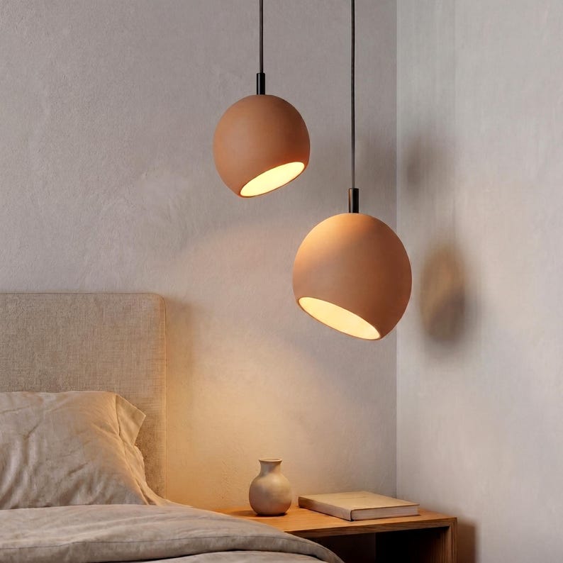 May include: Two terracotta-colored pendant lights with a spherical design hang from black cords. Each light has a tilted, open bottom revealing a warm, glowing interior. The lights are suspended above a bed and nightstand, suggesting a bedroom setting.