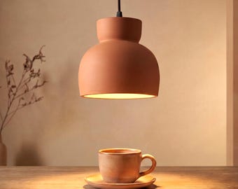 May include: A terracotta-coloured pendant light with a rounded dome shape hangs above a matching ceramic cup and saucer. The light fixture emits a warm glow, illuminating the cup and saucer set which rests on a wooden surface below the light.