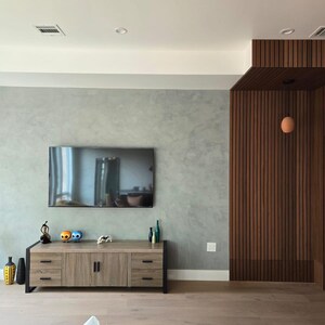 May include: A modern living room featuring a large flat-screen TV mounted on a gray wall. A wooden cabinet with black legs holds decorative items. The room has wood paneling and flooring, with a small plant in a red pot.