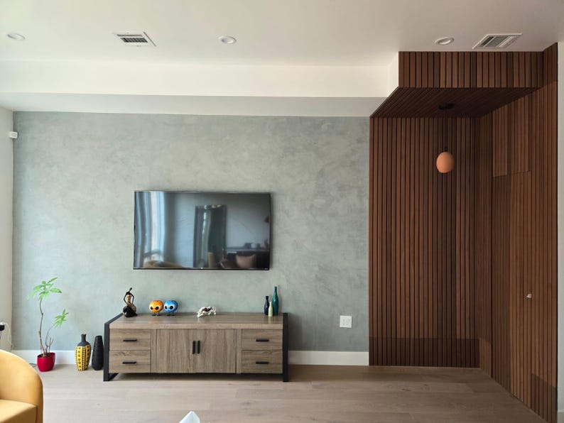 May include: A modern living room featuring a large flat-screen TV mounted on a gray wall. A wooden cabinet with black legs holds decorative items. The room has wood paneling and flooring, with a small plant in a red pot.