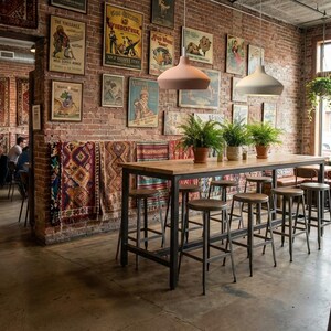 May include: A high wooden table with a metal frame surrounded by several metal bar stools in a room with exposed brick walls. The walls are decorated with framed vintage posters and hanging rugs. Two pendant lights hang above the table.