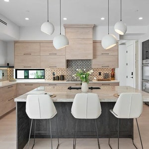 May include: Modern kitchen with light wood cabinetry, a dark gray island with a marble countertop, and white bar stools. White globe pendant lights hang above the island. A vase of white flowers sits on the island.