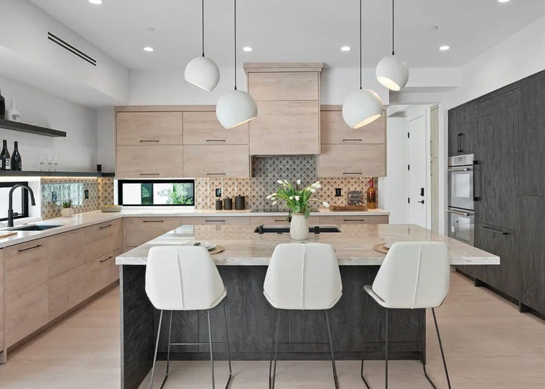 May include: Modern kitchen with light wood cabinetry, a dark gray island with a marble countertop, and white bar stools. White globe pendant lights hang above the island. A vase of white flowers sits on the island.
