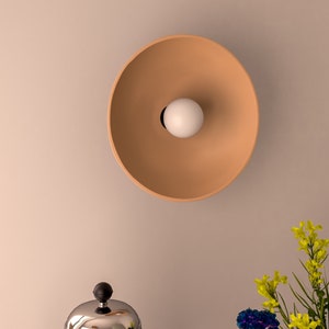 Könnte beinhalten: Eine Wandleuchte in Terrakottafarbe mit einem runden, flachen Schirm und einer weißen Glühbirne. Die Leuchte ist an einer weißen Wand montiert.