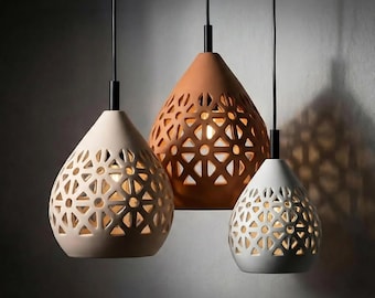 May include: Three pendant lights with a teardrop shape and intricate cut-out patterns. The lights are in cream, terracotta, and white, each suspended by a black cord. The cut-outs allow the warm light from within to shine through.
