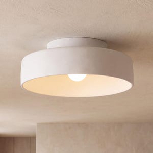 May include: A matte white flush mount ceiling light fixture with a wide, circular drum shape. The fixture features a smooth, solid exterior surface and a central light bulb that emits a soft, warm glow from within the open, shallow basin.