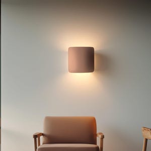 May include: A square, flush-mount wall sconce in a muted rose color is mounted on a light gray wall. The sconce emits a warm, inviting light, casting a soft glow downward. The design is minimalist and modern, suitable for various interior styles.