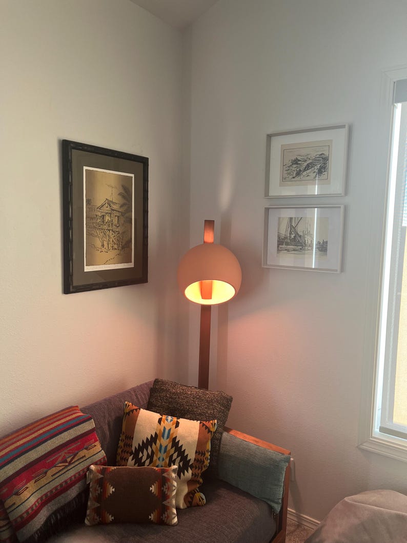 May include: A floor lamp with a spherical, matte peach-colored shade and a wooden stand, positioned next to a gray sofa adorned with a multi-colored striped throw blanket and two patterned accent pillows featuring geometric designs.