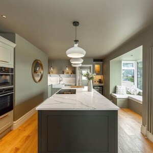 May include: A kitchen interior featuring a central island with a white marble countertop and dark gray base cabinetry. The space includes light green walls, light wood flooring, a built-in oven stack, three white pendant lights, and a window seat.