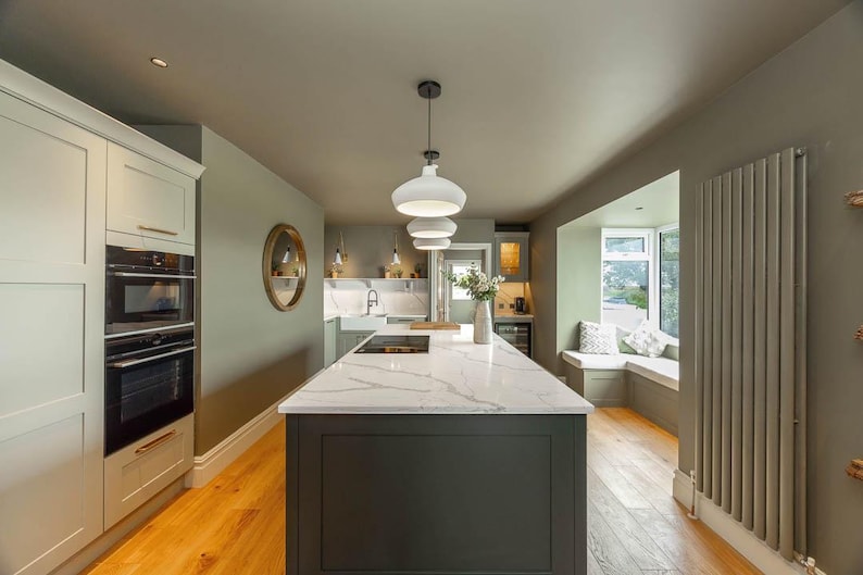 May include: A kitchen interior featuring a central island with a white marble countertop and dark gray base cabinetry. The space includes light green walls, light wood flooring, a built-in oven stack, three white pendant lights, and a window seat.