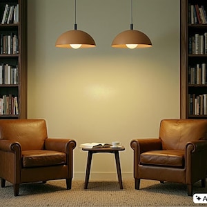 May include: Two brown leather armchairs face each other in a library setting, with a small table between them. Two pendant lights hang above. Bookshelves filled with books flank the chairs. A rug covers the floor.