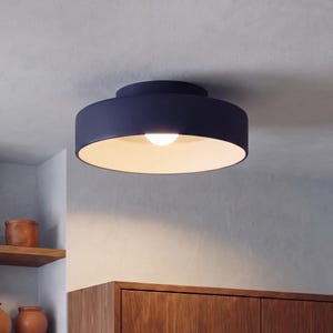 May include: A circular flush mount ceiling light fixture with a matte dark navy blue exterior finish and a smooth white interior surface. The fixture features a single central light bulb that emits a warm glow, illuminating the white underside.