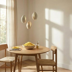 May include: A dining room scene featuring a round wooden table set for a meal. Three pendant lights hang above the table. Two wooden chairs with woven seats are positioned around the table. A bowl of pasta and a plate of food are on the table.