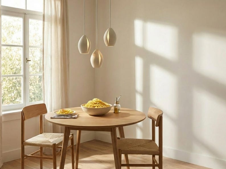 May include: A dining room scene featuring a round wooden table set for a meal. Three pendant lights hang above the table. Two wooden chairs with woven seats are positioned around the table. A bowl of pasta and a plate of food are on the table.