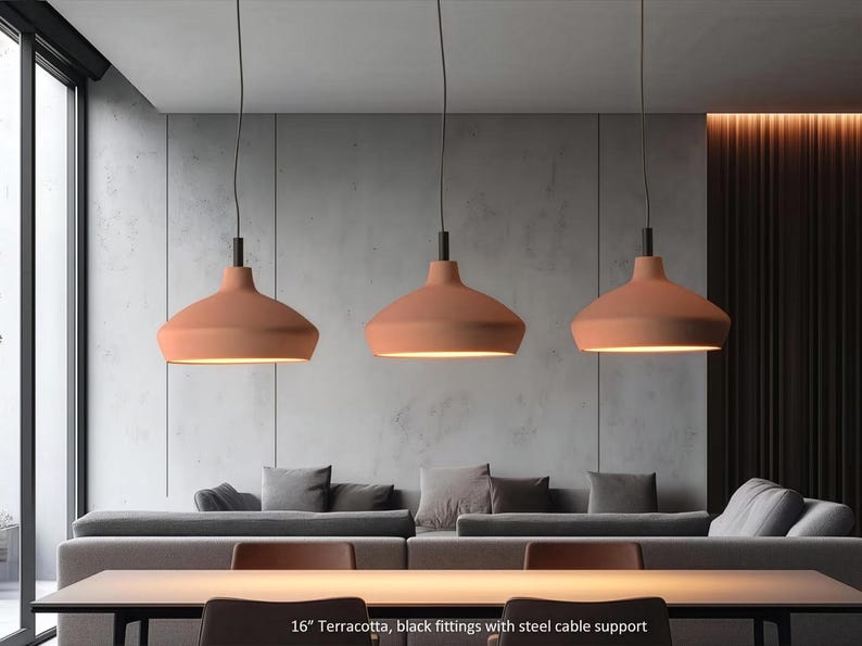 May include: Three 16-inch terracotta pendant lights with black fittings and steel cable supports. Each lamp features a wide, smooth, matte-finished dome shade that emits a warm downward glow, suspended from the ceiling by thin black cables.
