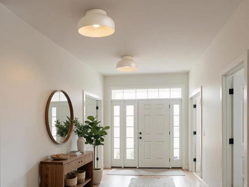 A bright, well-lit hallway with white walls and a light wood floor. Two white, dome-shaped ceiling lights illuminate the space. A wooden console table with a round mirror and potted plants adds a touch of warmth to the entryway.