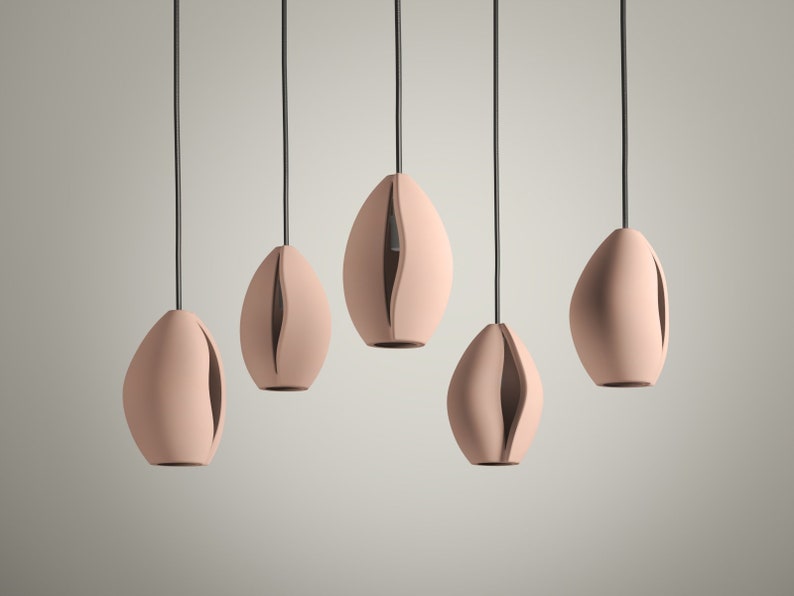 May include: Five modern pendant lights with a soft pink finish. Each light has an organic, oval shape with a vertical slit revealing the interior. The lights hang from black cords, creating a minimalist aesthetic.